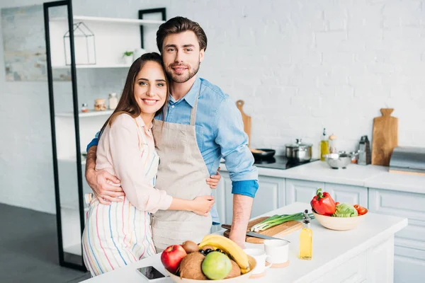 Casal em aventais abraçando e olhando para a câmera na cozinha — Fotografia de Stock