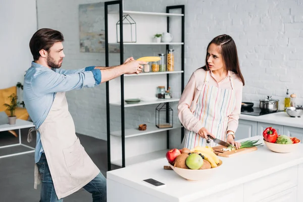 Petite amie couper des légumes et petit ami sérieux s'amuser avec pistolet à banane dans la cuisine — Photo de stock