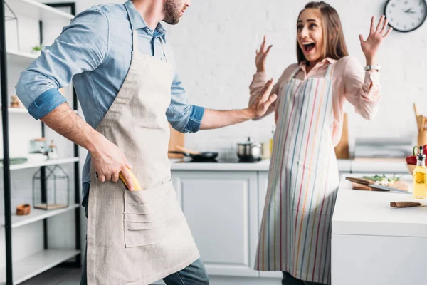 Excitada novia de pie con las manos arriba y novio sosteniendo pistola de plátano en la cocina - foto de stock