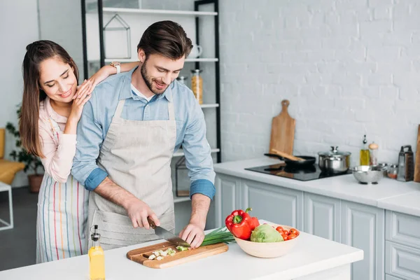 Novio cortar verduras y novia apoyándose en él en la cocina - foto de stock