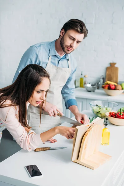 Хлопець ріже овочі і дівчина читає рецепт на кухні — стокове фото