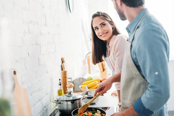 Fidanzato friggere verdure su padella in cucina e guardando sorridente fidanzata — Foto stock