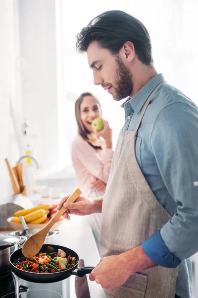 Boyfriend frying vegetables on frying pan in kitchen and girlfriend eating apple — Stock Photo