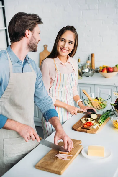 Sonriente pareja de cocinar y cortar verduras con carne en la cocina - foto de stock