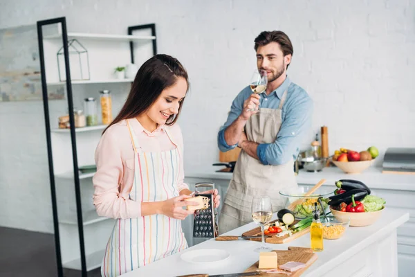 Sonriente novia rallar queso y novio beber vino en la cocina - foto de stock