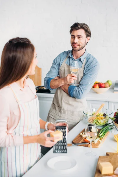 Girlfriend grating cheese and boyfriend drinking wine in kitchen — Stock Photo