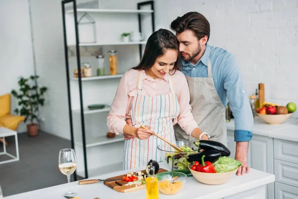 Novia cocinando y mezclando ensalada en cocina y novio abrazándola - foto de stock