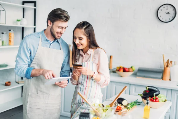 Feliz pareja de compras en línea con la tableta y tarjeta de crédito en la cocina - foto de stock