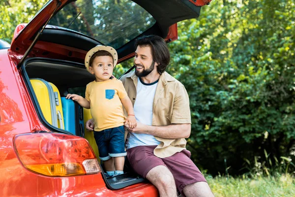 Sonriente padre e hijo sentado en el maletero del coche en el bosque - foto de stock