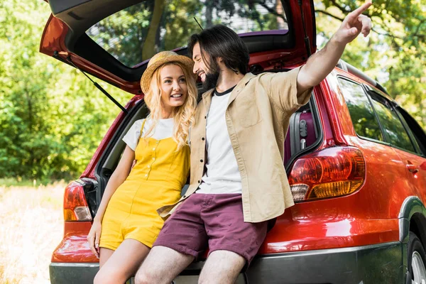 Boyfriend pointing on something to girlfriend while sitting on car trunk — Stock Photo