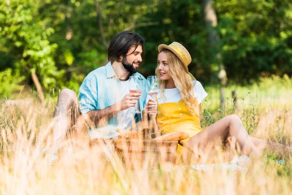 Pareja sentada en la hierba, sosteniendo vasos de vino y mirándose en el picnic - foto de stock