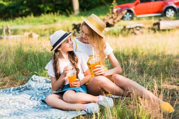 Мати і дочка сидять на ковдрі на пікніку, дивлячись один на одного і тримаючи склянки соку — стокове фото