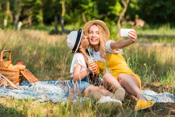 Daughter kissing mother while she taking selfie with smartphone at picnic — Stock Photo