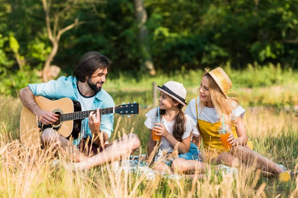 Padre feliz tocando la guitarra acústica para hija y esposa en el picnic - foto de stock