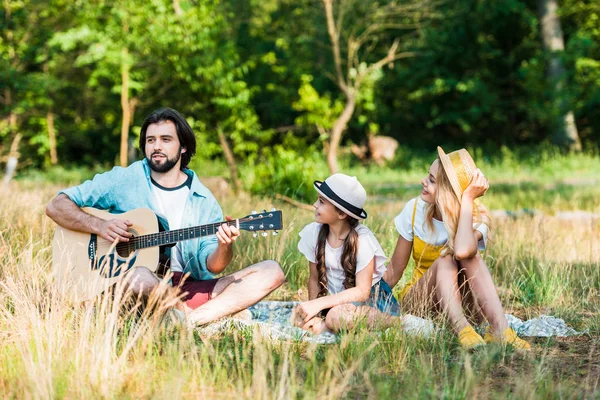 Padre tocando la guitarra acústica para hija y esposa en el picnic - foto de stock