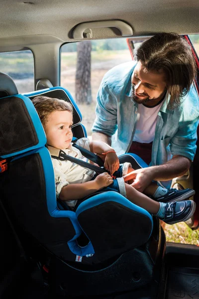 Улыбающийся отец крепит сына на безопасном сидении в машине — стоковое фото