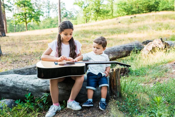 Siblings sitting on log at lawn with acoustic guitar — Stock Photo