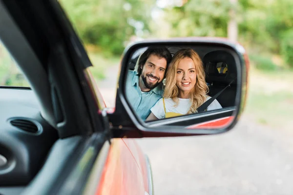 Sonriente pareja reflejándose en el espejo del coche y mirando a la cámara - foto de stock