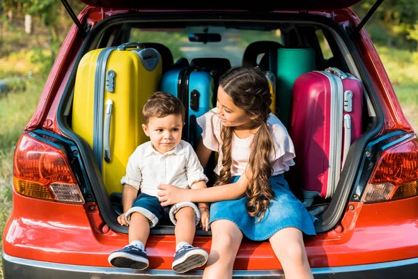 Adorable brother and sister sitting near travel bags in car trunk — Stock Photo