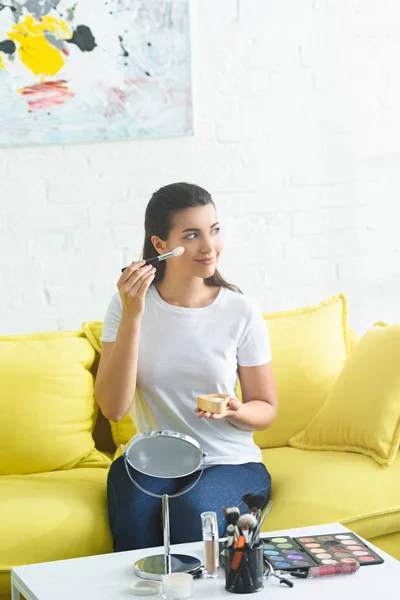 Retrato de la mujer mirando hacia otro lado y aplicando rubor facial mientras hace maquillaje en casa - foto de stock