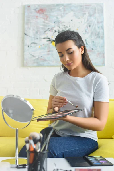 Porträt einer jungen Frau mit Make-up-Palette in den Händen, die Lidschatten aufträgt, während sie sich zu Hause auf dem Sofa schminkt — Stockfoto