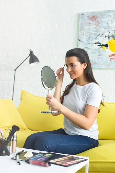 Вид сбоку на молодую женщину, смотрящую в зеркало и наносящую тени на глаза, делая макияж на диване дома — стоковое фото