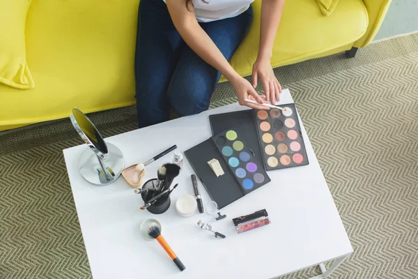Cropped shot of woman with makeup brush sitting on sofa at coffee table with cosmetics for makeup at home — Stock Photo