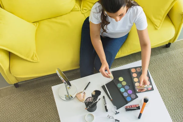 Vista aérea de la mujer sentada en el sofá en la mesa de café con cosméticos para el maquillaje en casa - foto de stock