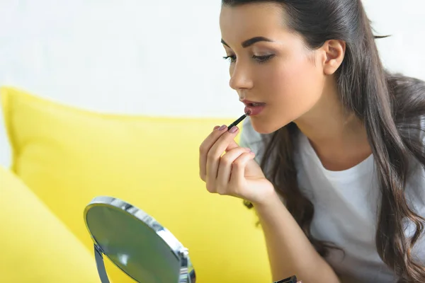 Vista lateral de la mujer joven aplicando lápiz labial mientras hace maquillaje en el sofá en casa - foto de stock