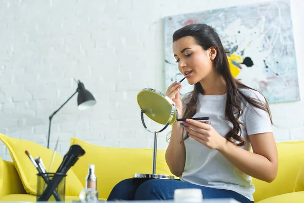 Молодая женщина наносит помаду, делая макияж на диване дома — стоковое фото