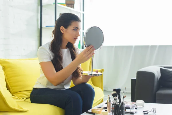 Вид сбоку молодой красивой женщины, смотрящей в зеркало, сидя на диване за журнальным столиком с косметикой дома — стоковое фото