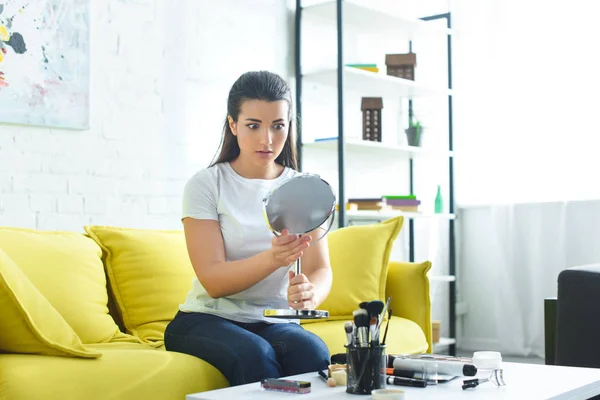 Портрет шокованої красивої жінки, дивлячись на дзеркало, сидячи на дивані за журнальним столиком з косметикою вдома — стокове фото
