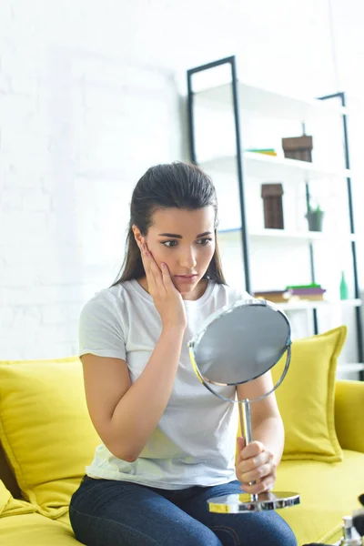 Portrait of shocked beautiful woman looking at mirror while sitting on sofa at coffee table with cosmetics at home — Stock Photo