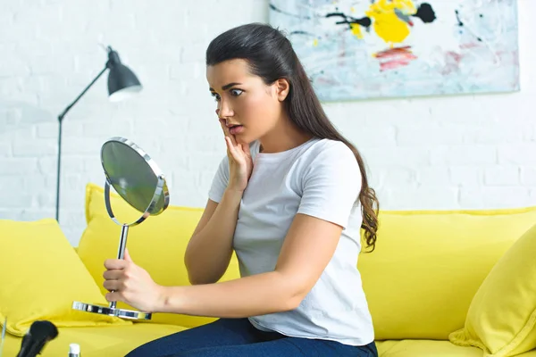 Side view of young shocked woman looking at mirror while sitting on sofa at coffee table with cosmetics at home — Stock Photo