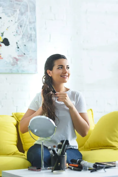 Ritratto di attraente donna sorridente spazzolare i capelli con pettine mentre seduto sul divano a casa — Foto stock