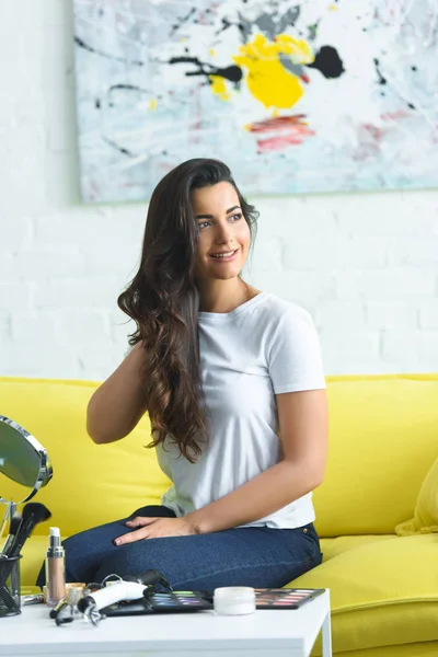 Красивая улыбающаяся женщина сидит на диване за журнальным столиком с зеркалом и косметикой дома — стоковое фото