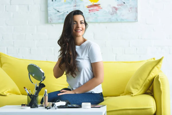 Schöne lächelnde Frau sitzt auf Sofa am Couchtisch mit Spiegel und Kosmetik zu Hause — Stockfoto