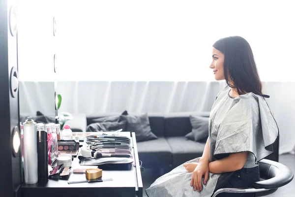Боковой вид молодой женщины, покрытой тканью, сидящей на стуле перед зеркалом и поверхностью с косметикой — стоковое фото
