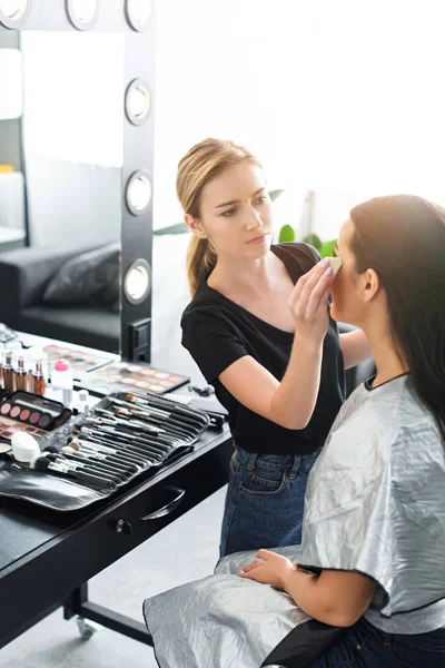 Vista lateral da bela jovem mulher recebendo maquiagem feita pelo artista de maquiagem focada — Fotografia de Stock
