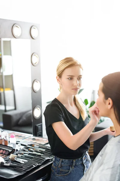 Selective focus of young woman getting makeup done by makeup artist — Stock Photo