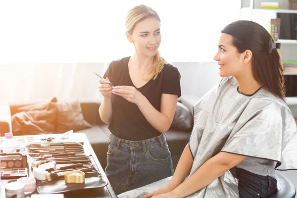 Vista lateral de artista de maquillaje sonriente con cosméticos y hermosa mujer joven cubierta con tela - foto de stock