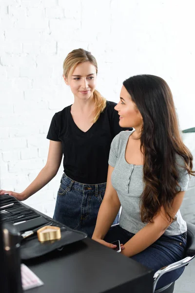 Smiling makeup artist and young woman with smartphone looking at each other — Stock Photo