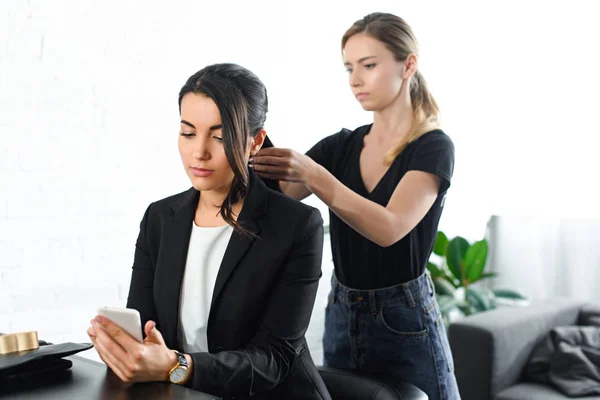 Hairstylist focado fazendo penteado enquanto empresária em terno usando smartphone — Fotografia de Stock