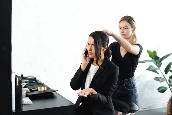 Coiffeur concentré faisant coiffure tandis que la femme d'affaires en costume parler sur smartphone — Photo de stock