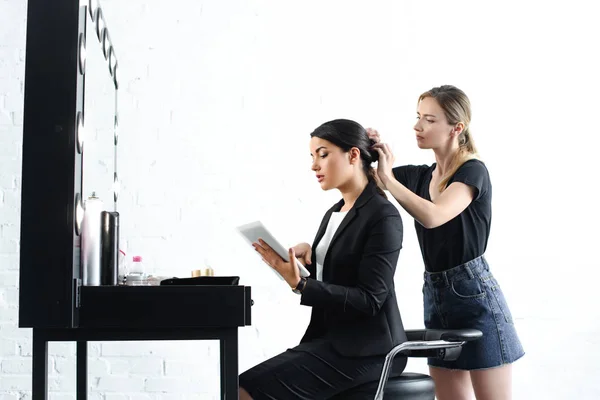 Vista laterale del parrucchiere facendo acconciatura mentre la donna d'affari in giacca e cravatta utilizzando tablet digitale — Foto stock