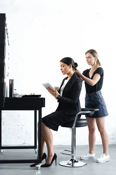 Vista lateral del estilista haciendo peinado mientras que la mujer de negocios en traje usando tableta digital - foto de stock