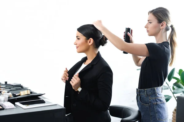 Vista lateral de la mujer de negocios consiguiendo el peinado fijado con el aerosol del pelo por el estilista - foto de stock