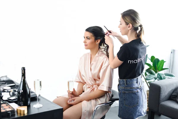 Young hairstylist doing hairstyle for bride in silk bathrobe with glass of champagne — Stock Photo