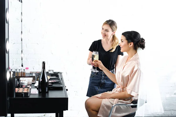 Maquillaje sonriente artista y novia en albornoz de seda con velo tintineo copas de champán - foto de stock