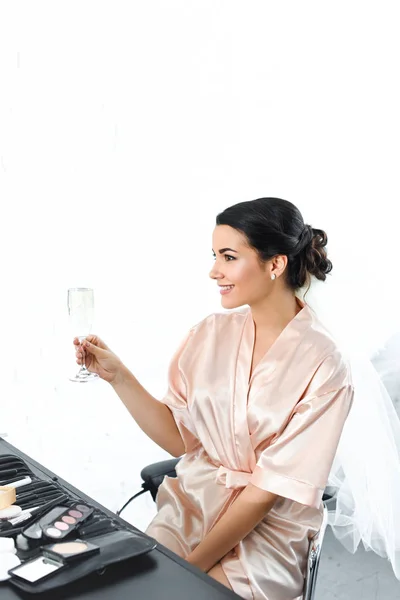 Side view of smiling bride in silk bathrobe and veil with glass of champagne sitting at tabletop with cosmetics — Stock Photo